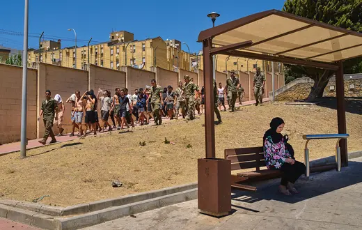 Mieszkanka Ceuty na przystanku autobusowym, za nią eskortowani przez żołnierzy nielegalni migranci, którzy w ostatnich dniach lipca wdarli się na teren hiszpańskiej eksklawy w Afryce | Fot. Bernat Armangue/Associated Press/East News